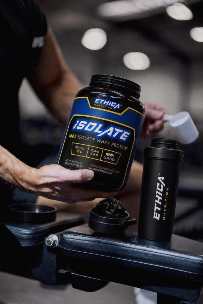 Athlete holding an Ethica Isolate whey protein tub and scooping powder beside an Ethica Nutrition shaker on a gym bench